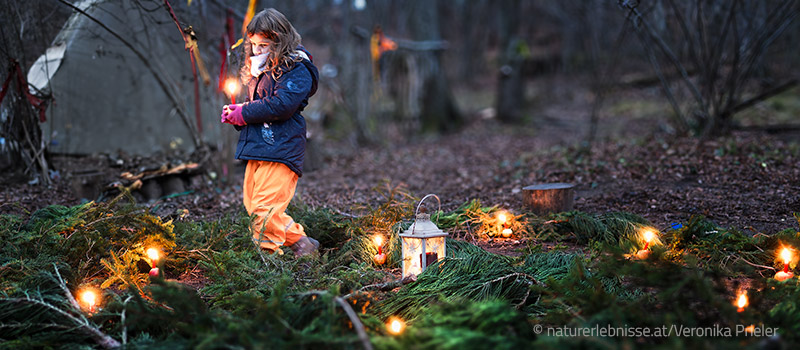 Adventliches Lichterfest im Wald: Kind geht achtsam durch Lichterspirale aus Reisig und Kerzen in der Dämmerung