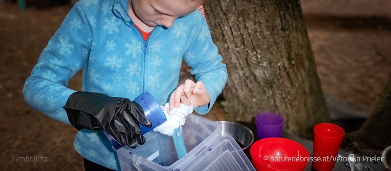 Ein Kind, im Wald, mit schwarzen Schutzhandschuhen gießt konzentriert eine farbige Flüssigkeit auf ein Tuch über einem Kunststoffbehälter – Titelbild für den Kinder-Workshop „Forscher-Action: Vulkan, Raketen & Wow-Experimente!"